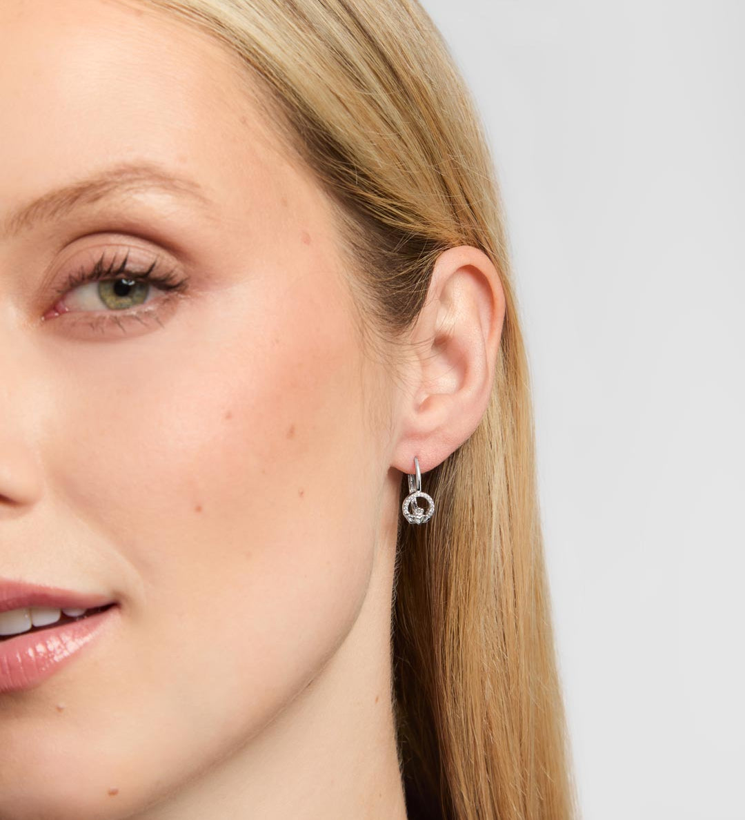 Close-up of a woman's ear wearing a delicate silver Claddagh hoop earring with a small dangling circular charm, against a light grey background.