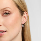 Close-up of a woman's ear wearing a delicate silver Claddagh hoop earring with a small dangling circular charm, against a light grey background.