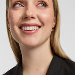 Close-up of a smiling woman looking upward, wearing small silver hoop earrings featuring Claddagh symbol and a black top against a light grey background.