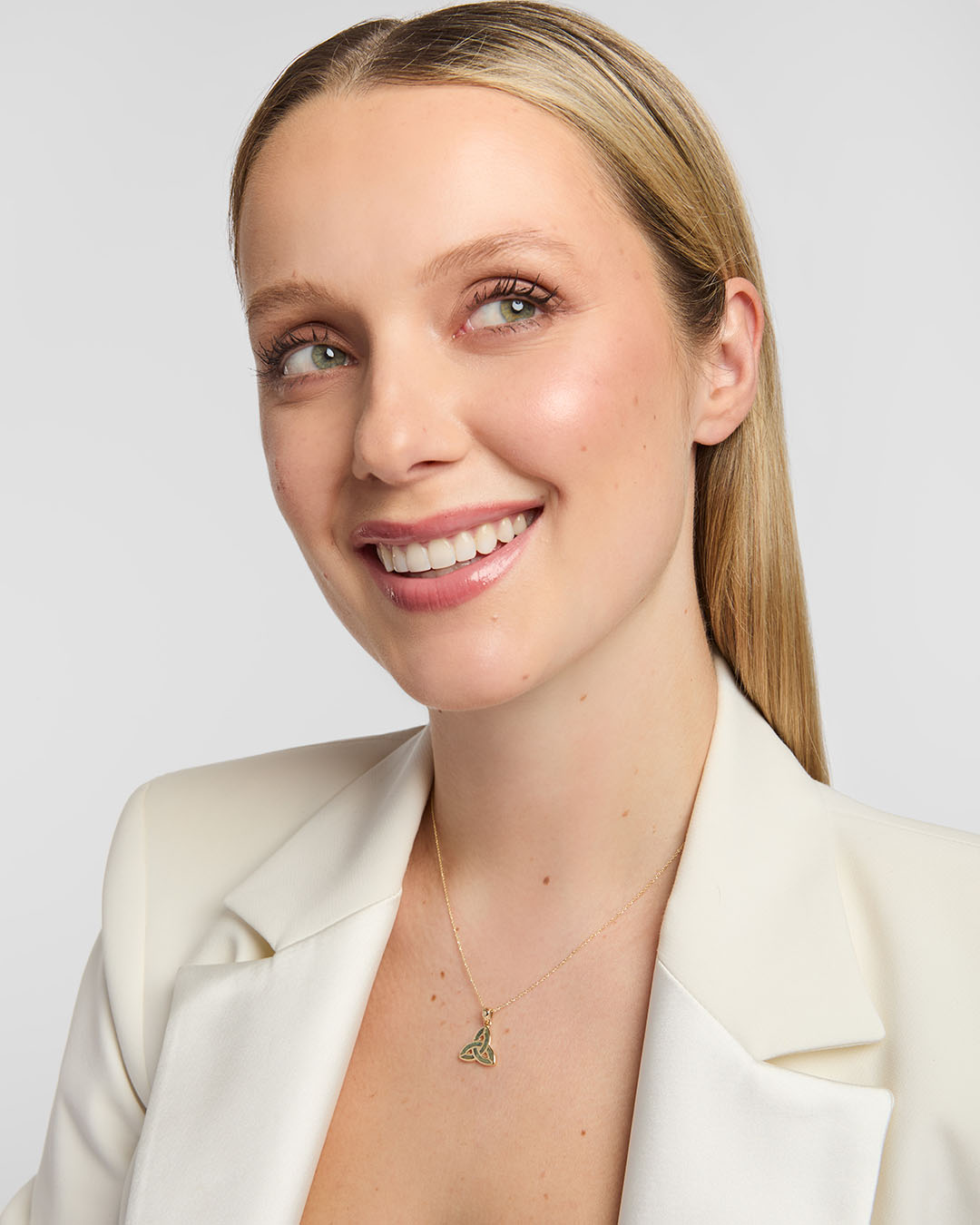A smiling blonde woman in a cream blazer wearing a delicate Solvar Connemara marble Trinity Knot necklace in gold. photographed against a grey studio background.