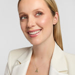A smiling blonde woman in a cream blazer wearing a delicate Solvar Connemara marble Trinity Knot necklace in gold. photographed against a grey studio background.