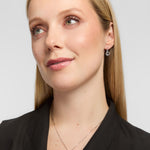 A close-up portrait of a woman wearing a delicate silver Claddagh pendant necklace and matching hoop earrings against a light grey background.
