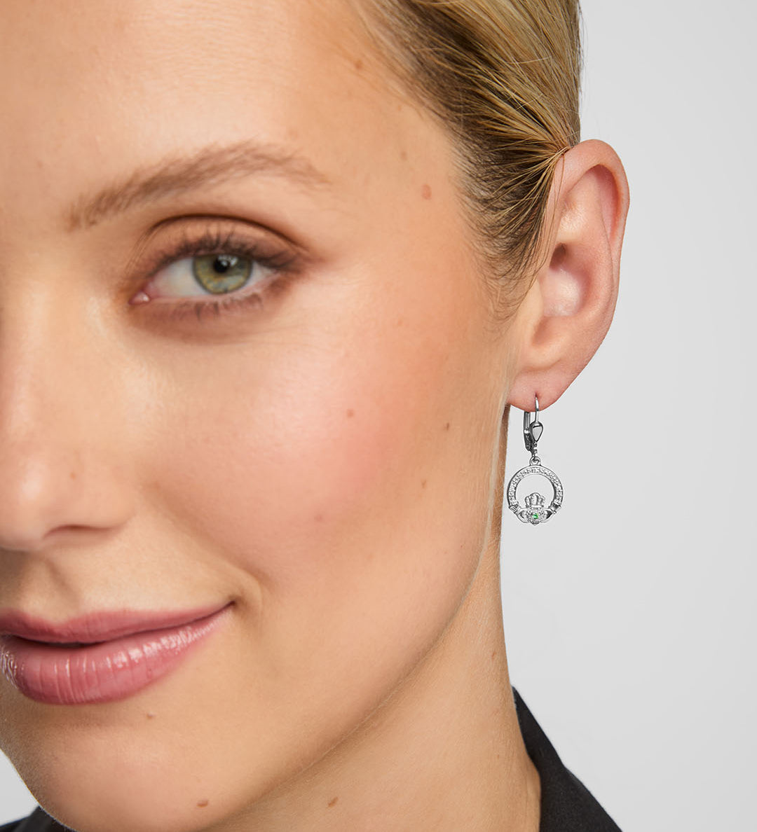 close-up image of female model wearing green crystal centre Claddagh drop earrings against grey background
