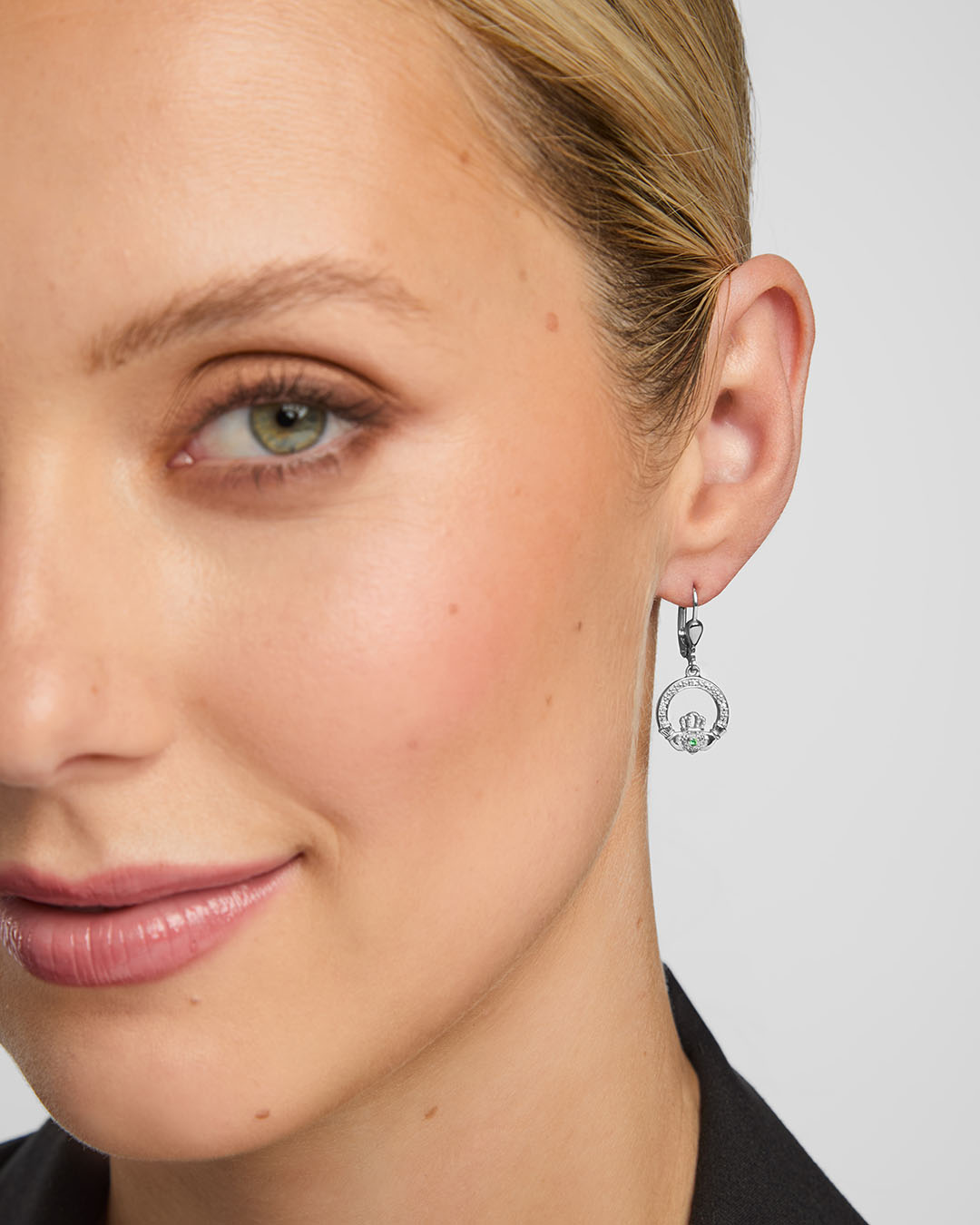 close-up image of female model wearing green crystal centre Claddagh drop earrings against grey background