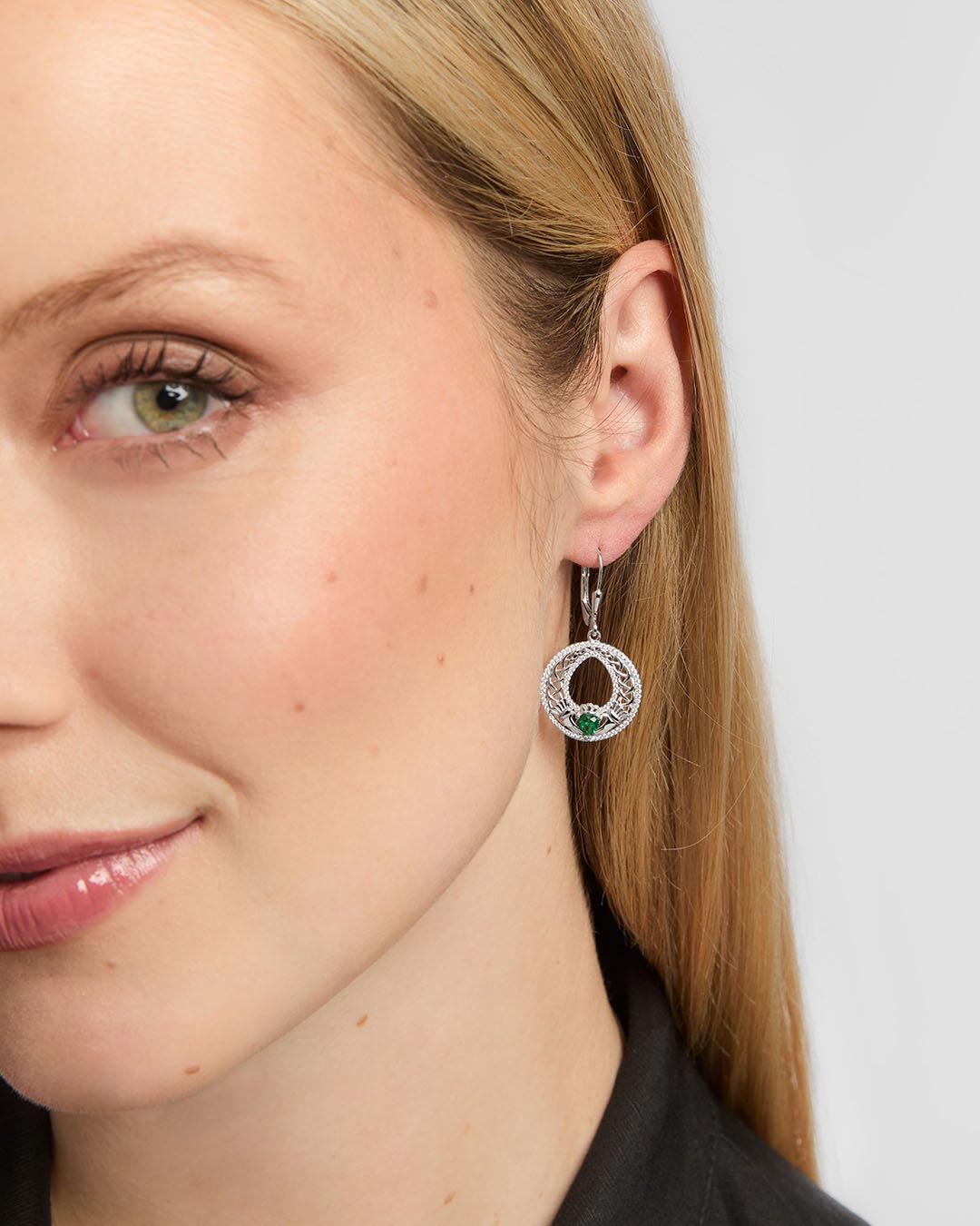 A close-up of a woman's ear and side profile wearing a silver Celtic Claddagh Drop earrings with a circular halo design and a green teardrop-shaped stone at the centre.