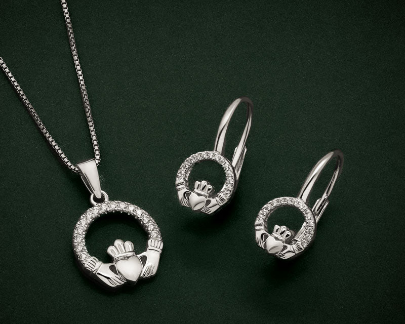 Silver-tone jewellery set featuring a Claddagh pendant necklace and matching hoop earrings, displayed on a dark green background.