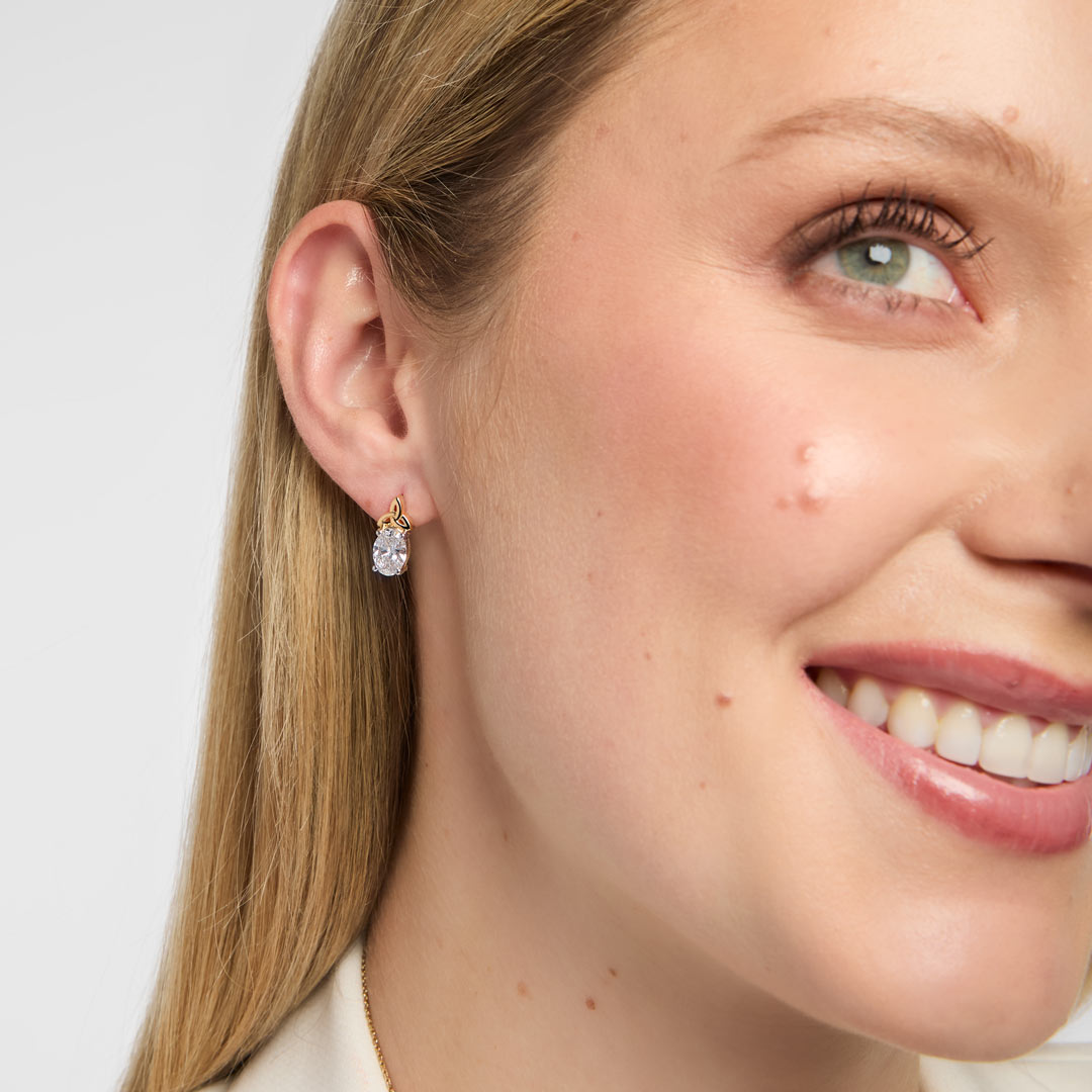 A close-up of a woman's wearing a small gold Trinity Knot stud earrings with a clear teardrop-shaped Lab diamond, set against a light grey background.