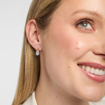 A close-up of a woman's wearing a small gold Trinity Knot stud earrings with a clear teardrop-shaped Lab diamond, set against a light grey background.