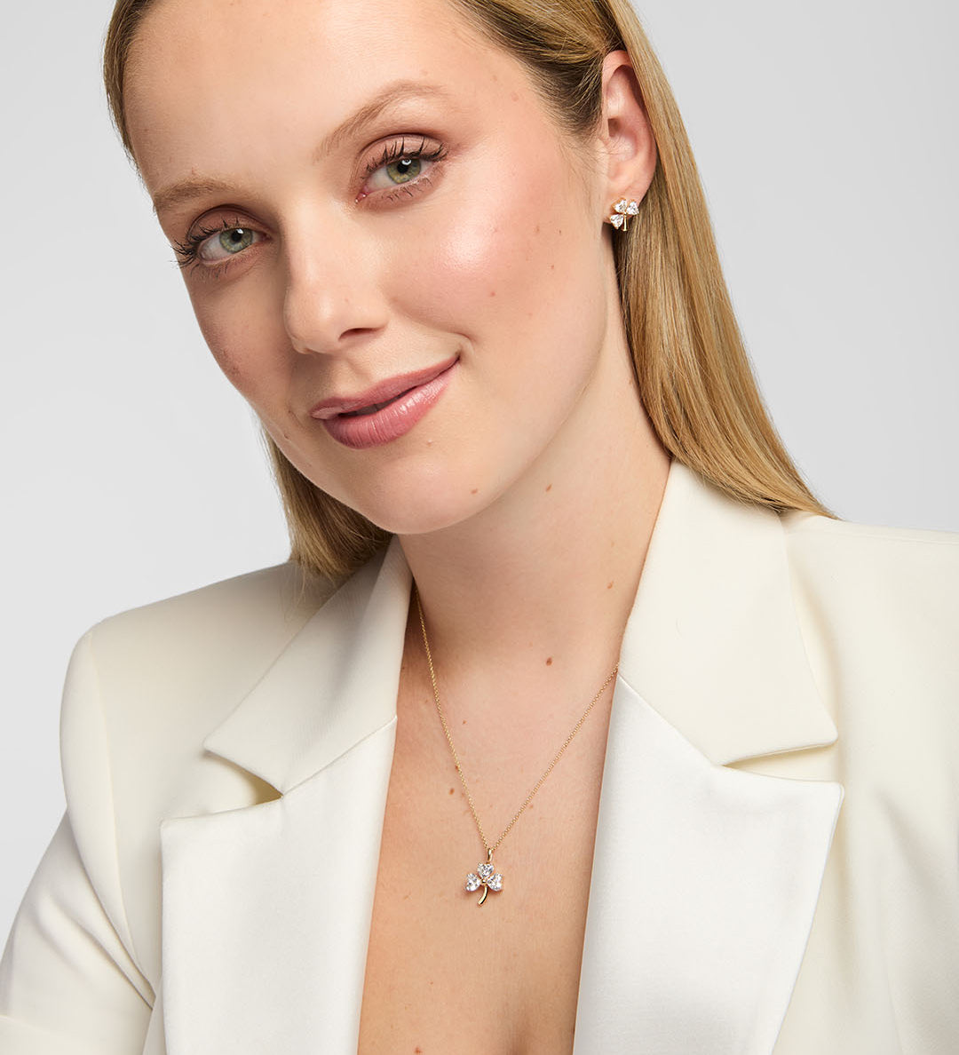 A close-up of a woman wearing a delicate lab-diamond gold necklace with a small shamrock shaped pendant and matching stud earrings, styled with a cream-colored blazer against a light grey background.