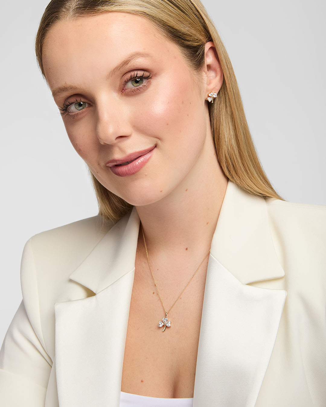 A close-up of a woman wearing a delicate lab-diamond gold necklace with a small shamrock shaped pendant and matching stud earrings, styled with a cream-colored blazer against a light grey background.