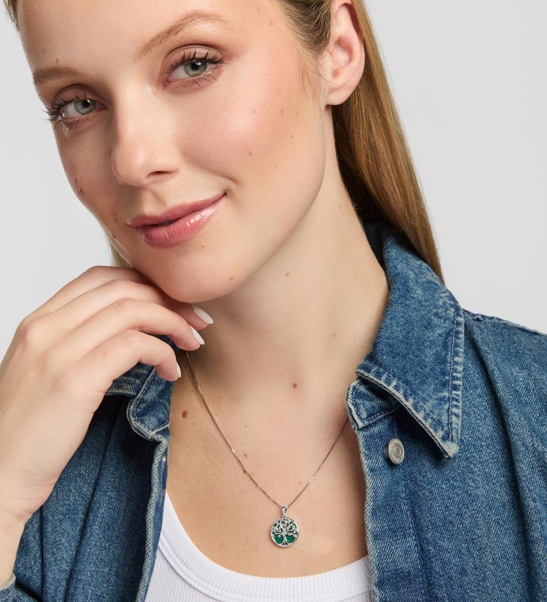 A close-up of a woman wearing a delicate silver Celtic necklace with a circular tree of life pendant featuring green malachite accents, styled with a blue denim shirt over a white top.