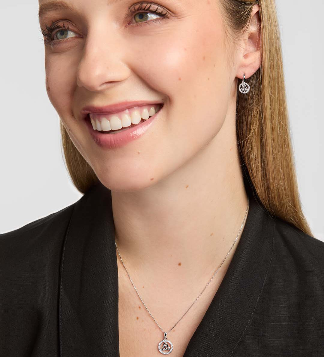 A close-up of a smiling woman wearing a delicate silver Celtic necklace with a circular Trinity Knot pendant and matching stud earrings against a light grey background.