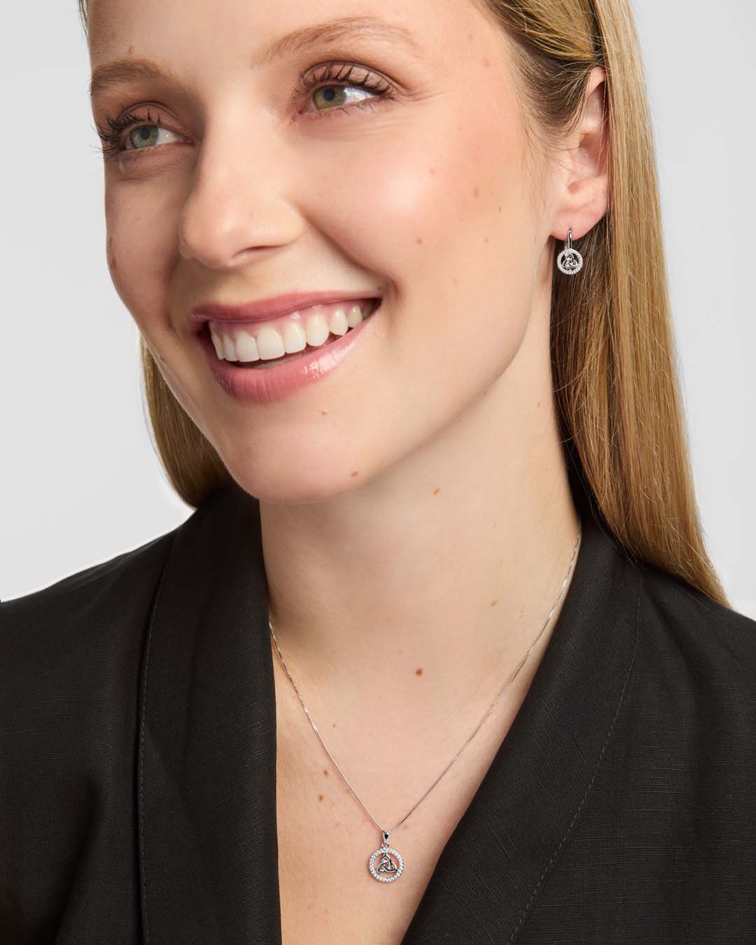 A close-up of a smiling woman wearing a delicate silver Celtic necklace with a circular Trinity Knot pendant and matching stud earrings against a light grey background.