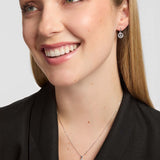 A close-up of a smiling woman wearing a delicate silver Celtic necklace with a circular Trinity Knot pendant and matching stud earrings against a light grey background.