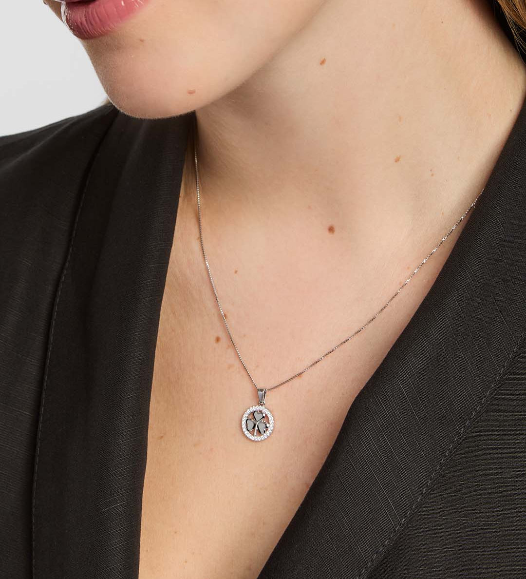 A close-up of a model wearing a delicate silver chain necklace with a circular pendant featuring a shamrock design surrounded by a clear stone halo.