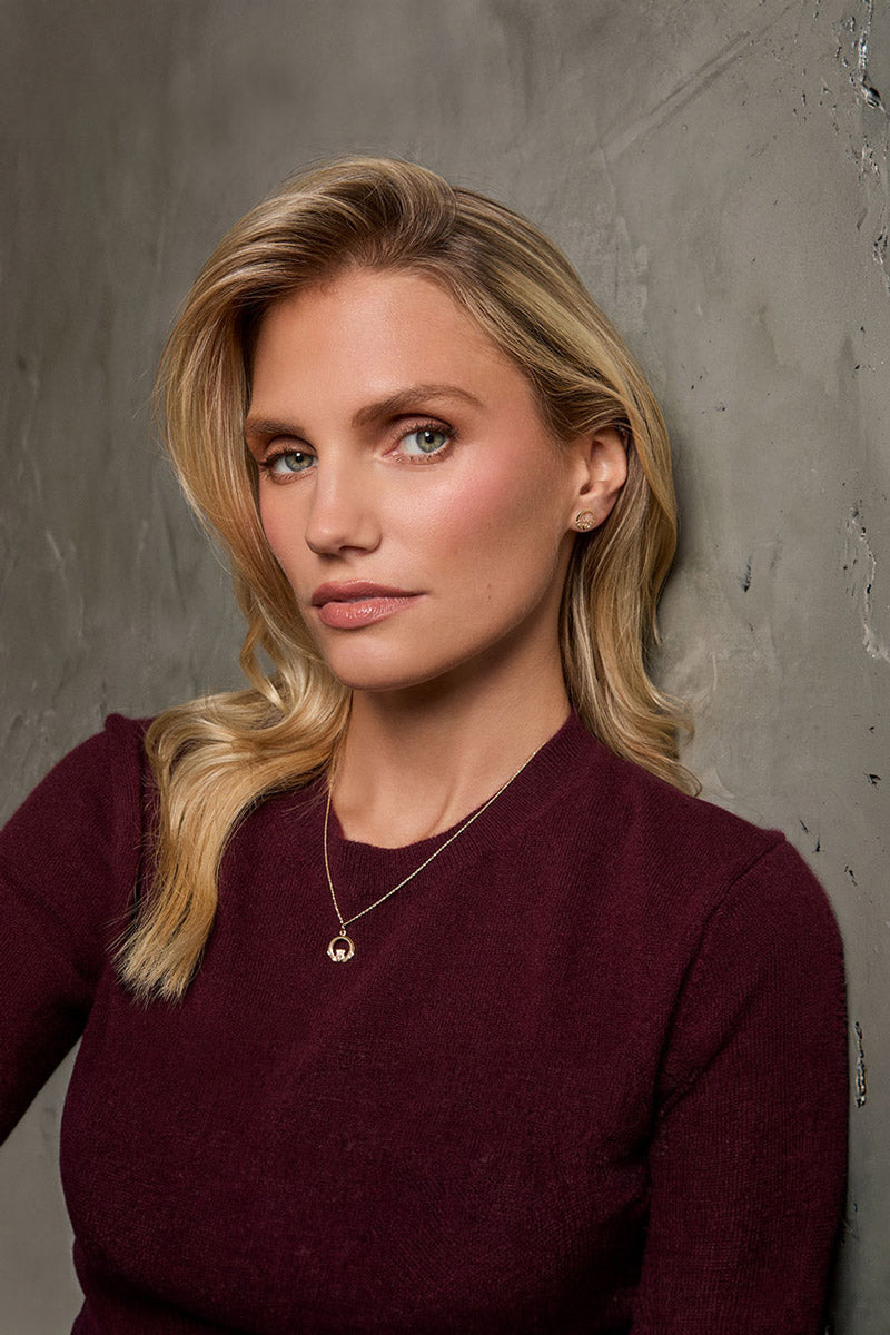 Model wearing Solvar gold Claddagh earrings with a classic plain gold Claddagh pendant necklace posing against a textured grey concrete wall.