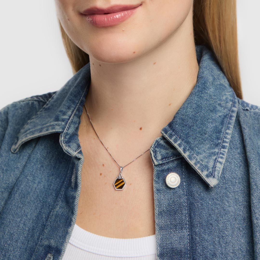 A close-up of a female model wearing a silver necklace with a small hexagonal pendant featuring a black and orange tiger eye striped pattern, styled with a blue denim shirt.