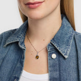A close-up of a female model wearing a silver necklace with a small hexagonal pendant featuring a black and orange tiger eye striped pattern, styled with a blue denim shirt.