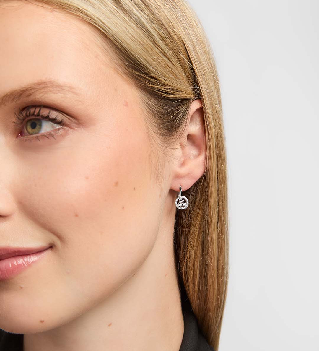 A close-up side profile of a woman wearing a small silver hoop earring with a Trinity Knot centre surrounded by sparkling clear stones.